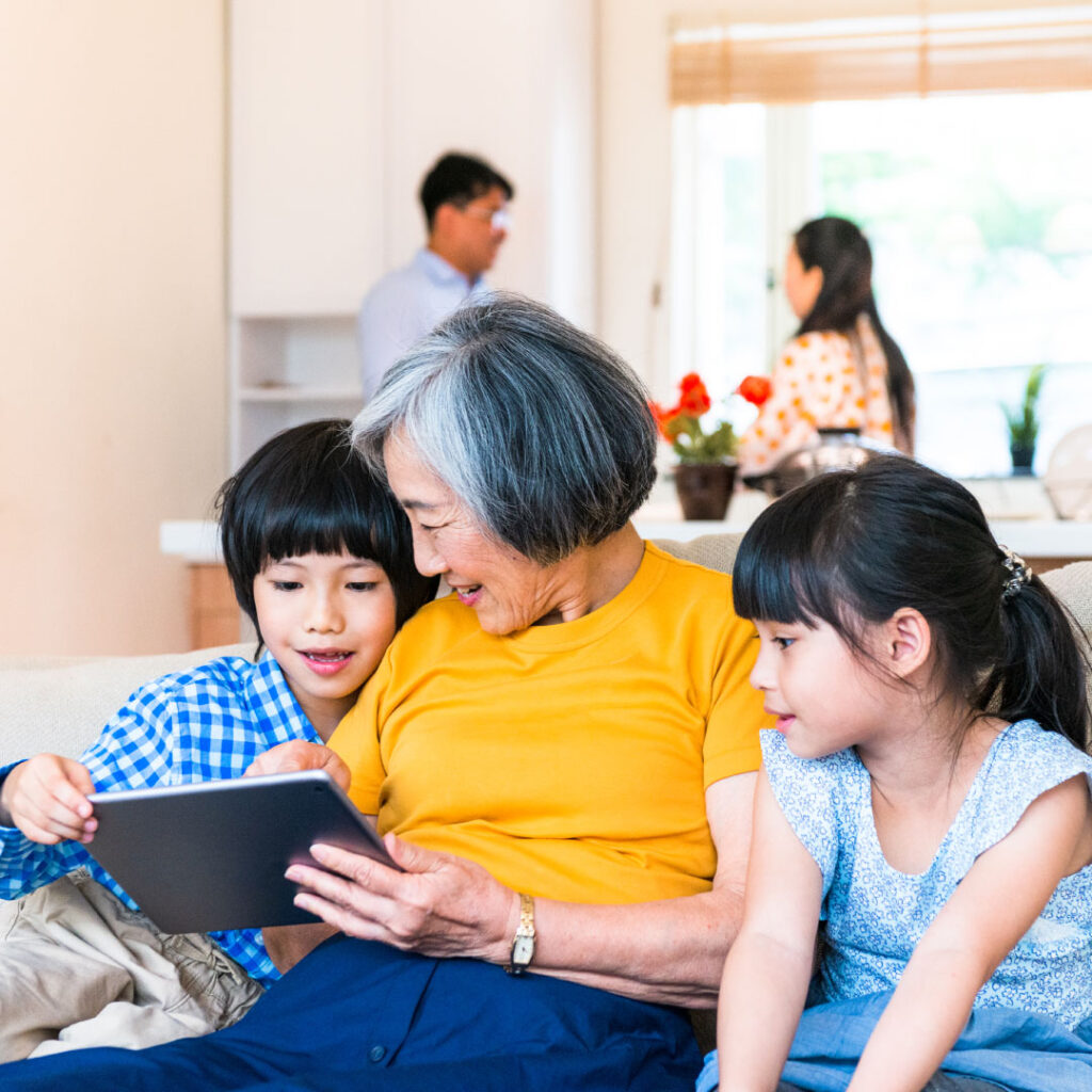 Senior woman with 2 young kids on couch. She is showing them something on a tablet with 2 young adults in the background.