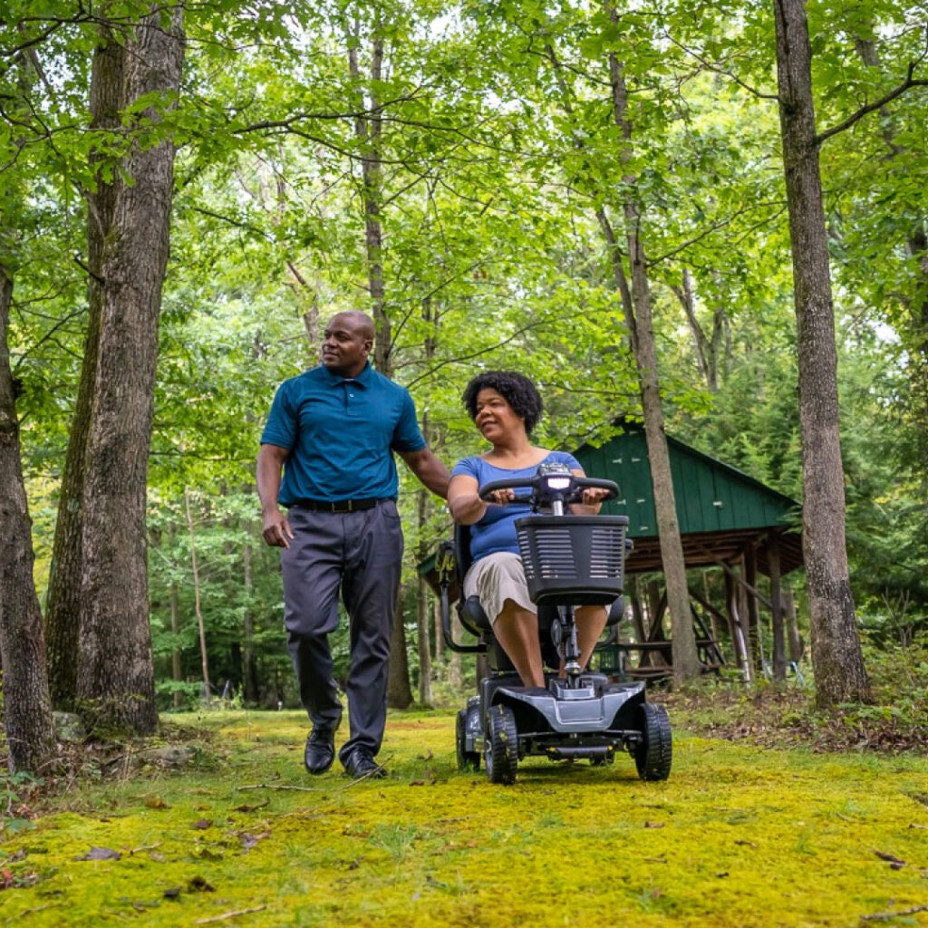 Woman on a mobility scooter with man to her right on a bath in the woods.