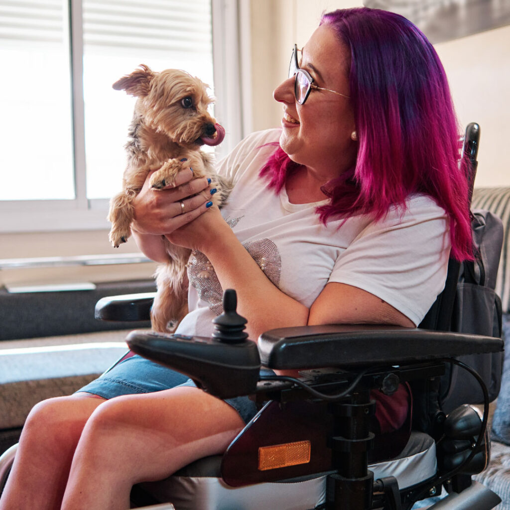 Young woman in a power wheelchair in the living room with a small brown and black dog on her lap.