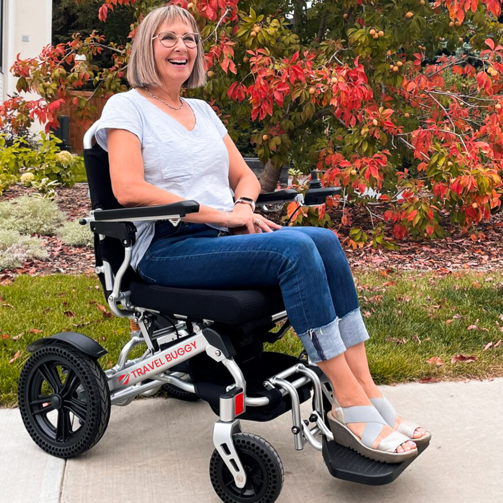 Woman seated in Travel Buggy Vista power wheelchair on sidewalk in front of red flowering shrub
