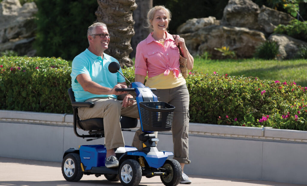 Man using 4-wheeled blue mobility scooter on summer day with woman walking on side