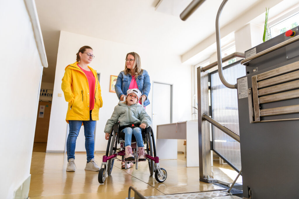 Mother and young woman with a young child living with cerebral palsy using manual wheelchair and waiting to access a vertical platform lift within a facility.