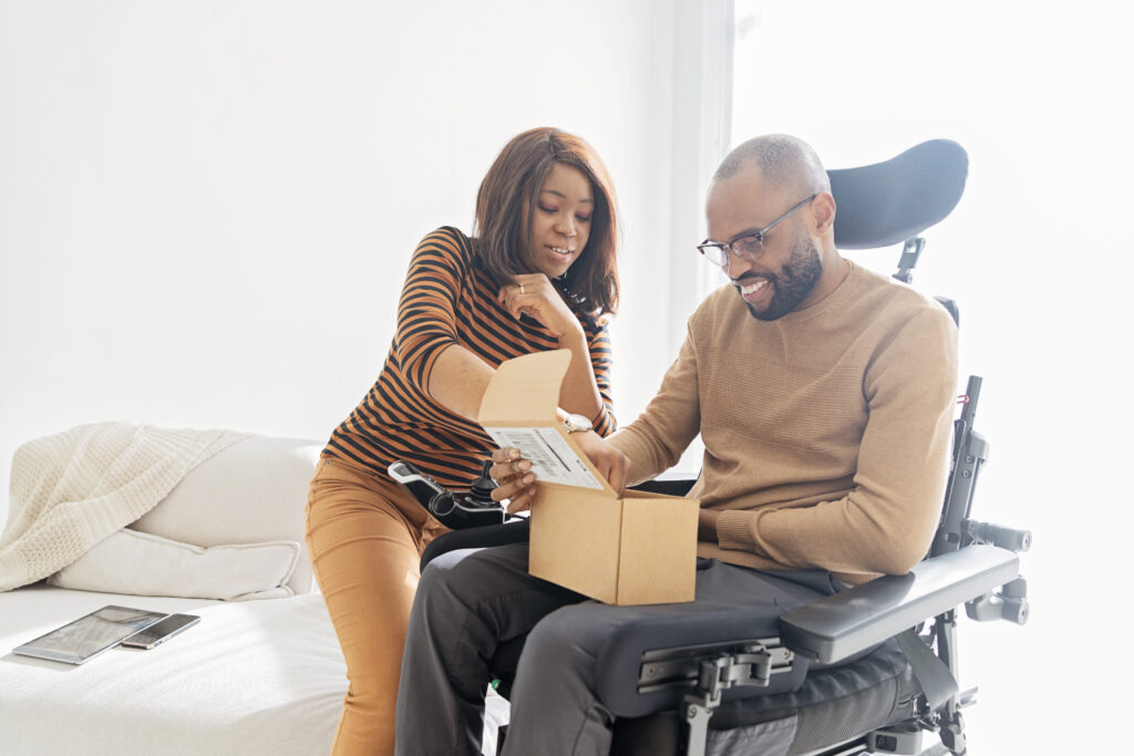 Side view of man in a power wheelchair with a woman hovering over a sofa next to him while he is opening a package