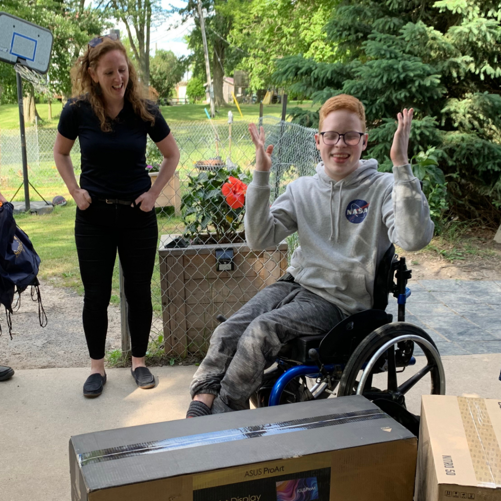 Shine Foundation dream recipient Corbett seated in a manual wheelchair with his hands pointed upwards in excitement. A box is pictured in front of him and a representative standing in background.