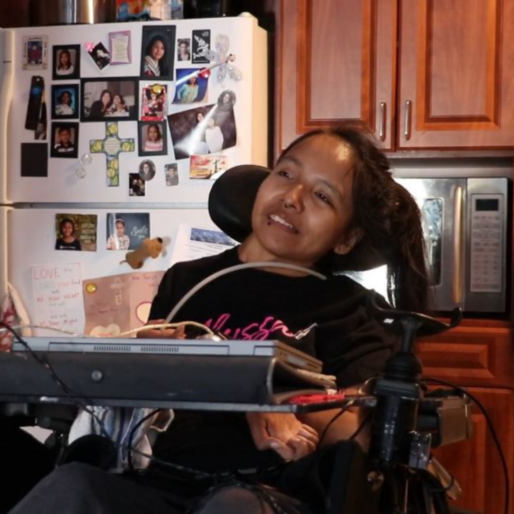 Motion client Maggie Sofea seated in her power wheelchair with front tray pictured in front of a white fridge and medium coloured wood cabinets in her kitchen