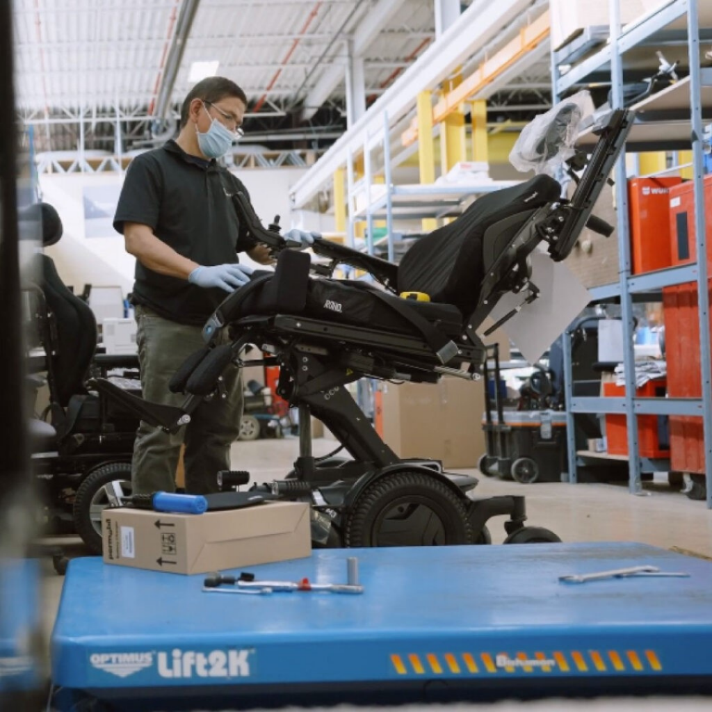 Motion technician repairing a power wheelchair with power positioning in a warehouse setting.