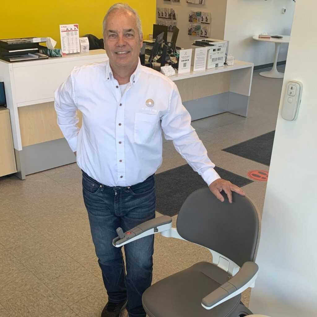 Dave V., Motion Representative in a Motion showroom standing and smiling with his hand resting on the top of a stairlift chair back. 