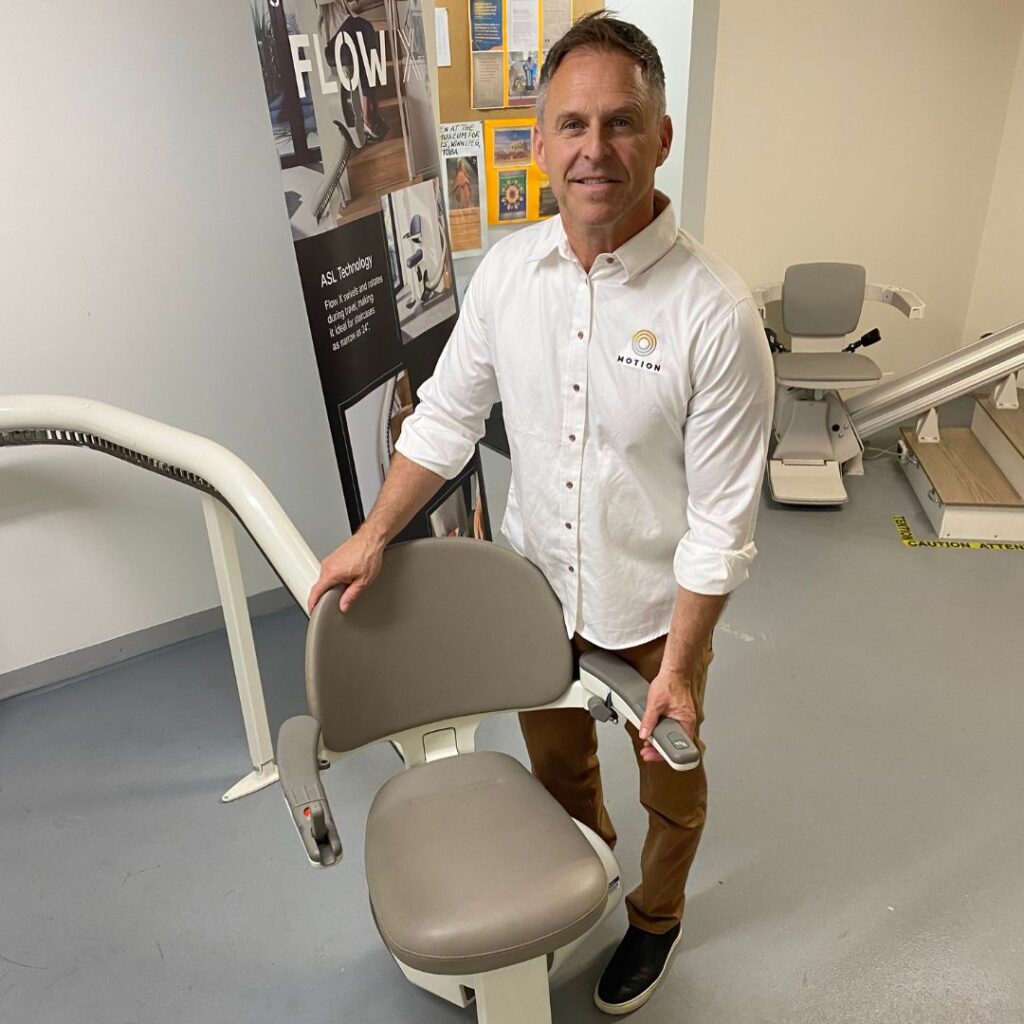 Motion Home Accessibility Specialist Thomas standing to the right of a stairlift seat and rail in a Motion showroom. Thomas is smiling and wearing a Motion-branded uniform shirt. 