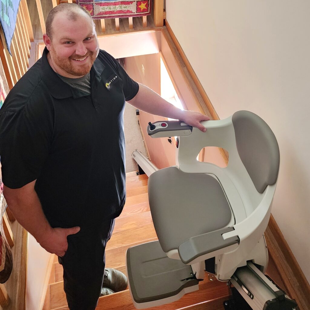 Motion Home Accessibility Specialist Paul standing at the top of a light wood staircase on the left of a stairlift seat and rail. Paul is smiling and wearing a Motion-branded uniform shirt. 