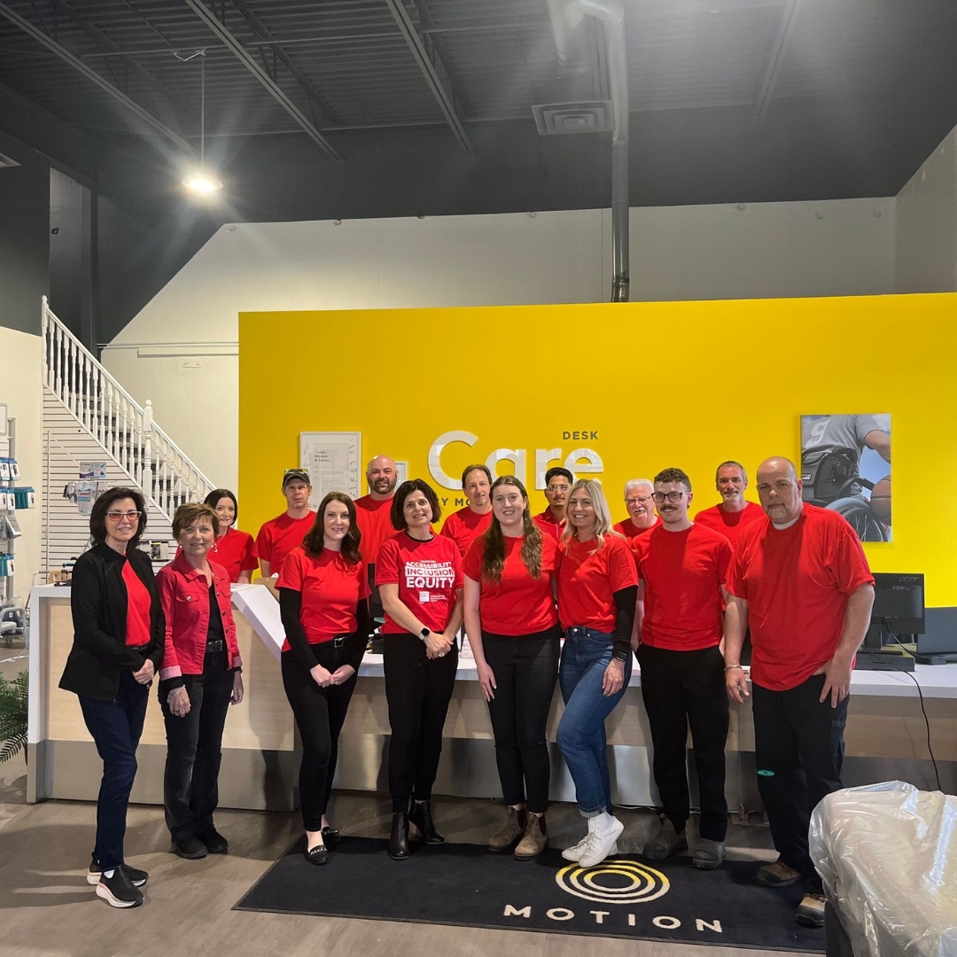 Motion CEO Sue Gilpin pictured in the centre of a large group of Motion employees wearing red for Red Shirt Day. The group is in a Motion showroom standing in front of a yellow Care Desk wall.