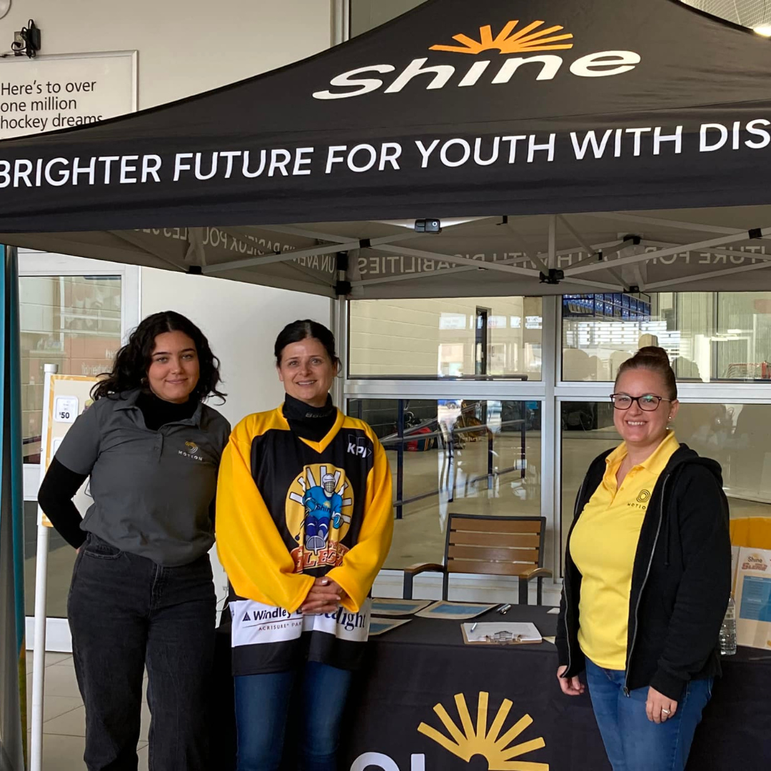 Motion CEO Sue Gilpin wearing a hockey jersey is standing in the middle of two Motion employees wearing Motion-branded uniforms. The group is standing in front of a Shine Foundation booth with tent and table.