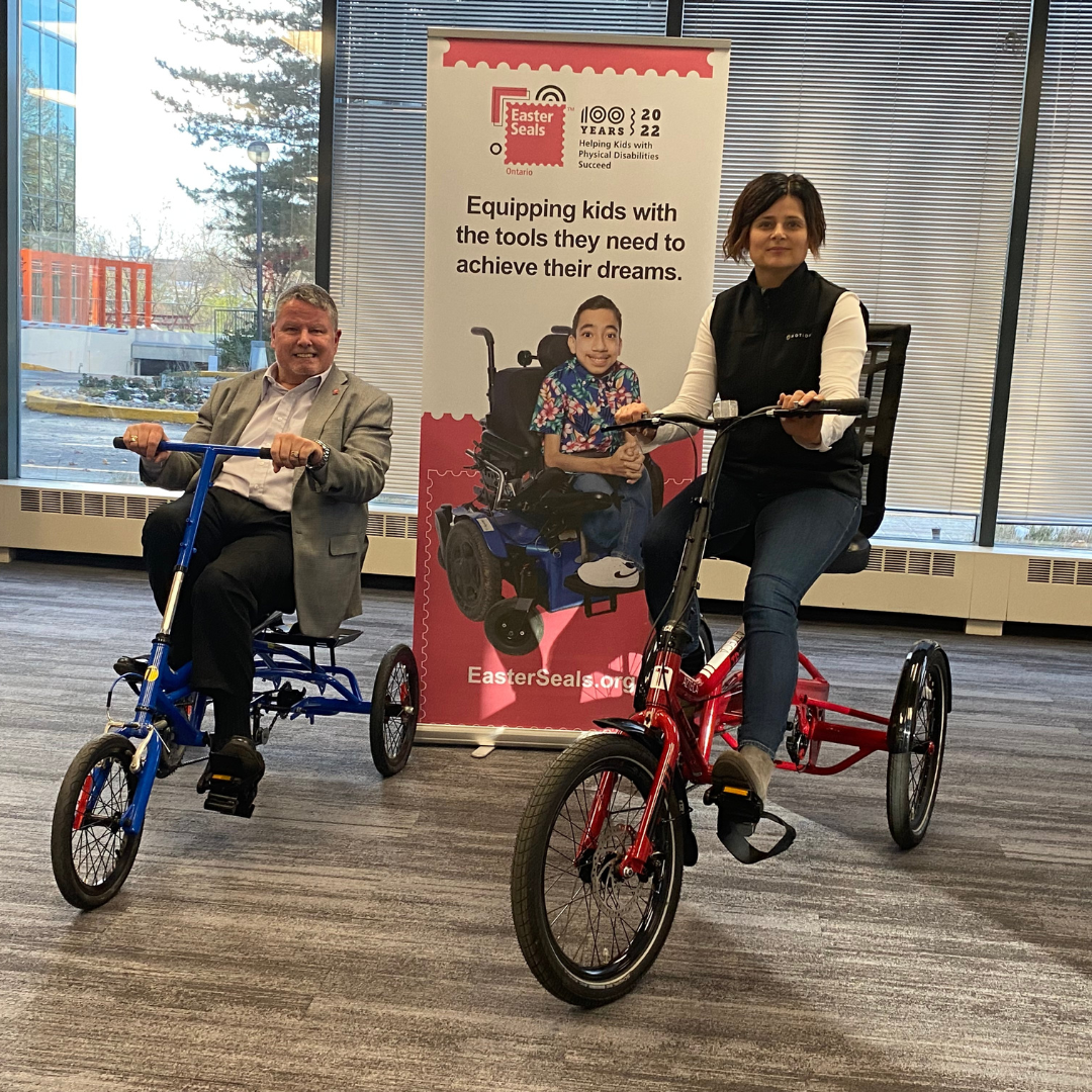 Motion CEO Sue Gilpin pictured with the Easter Seals Ontario CEO. Both are seated in adaptive bikes and there is an Easter Seals retractable banner behind them.