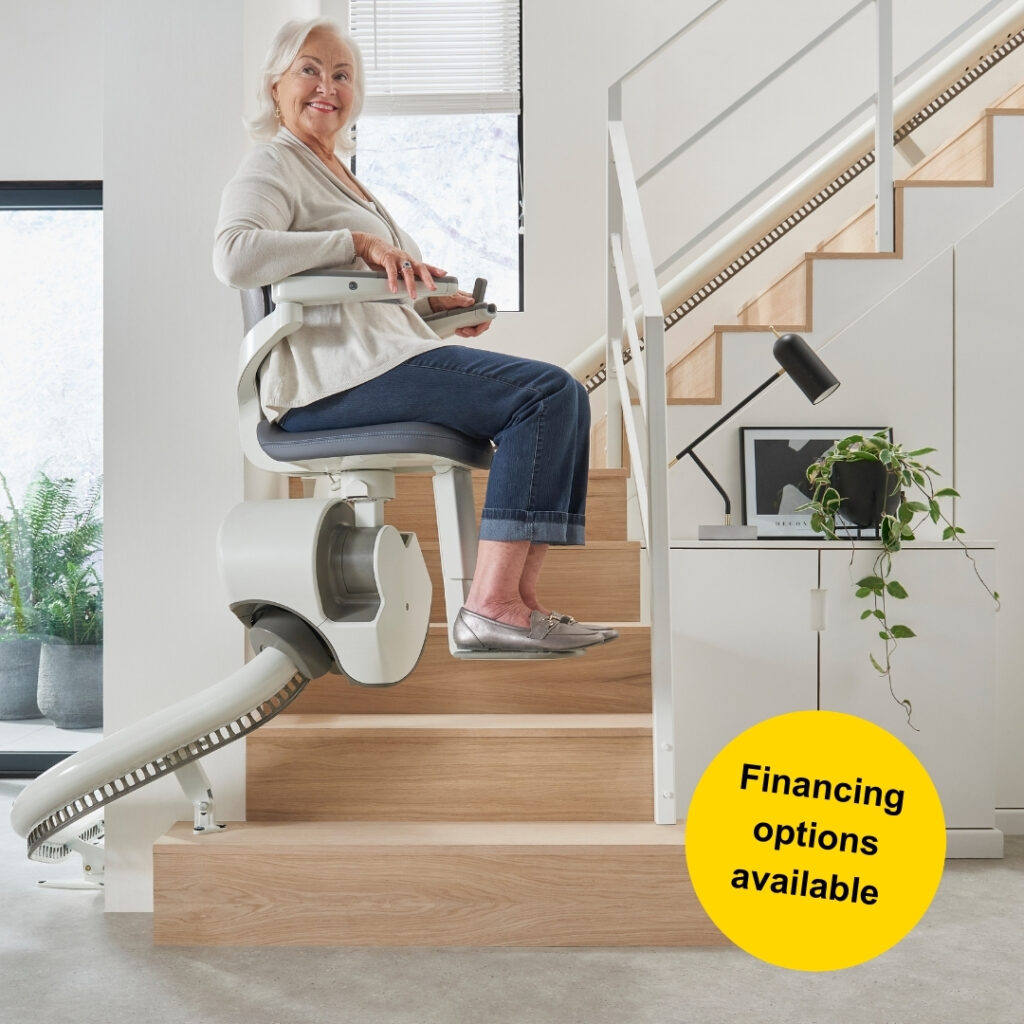 Senior woman pictured seated on stairlift chair at the bottom of a curved staircase. A round yellow sticker is in the bottom right corner and reads financing options available. 