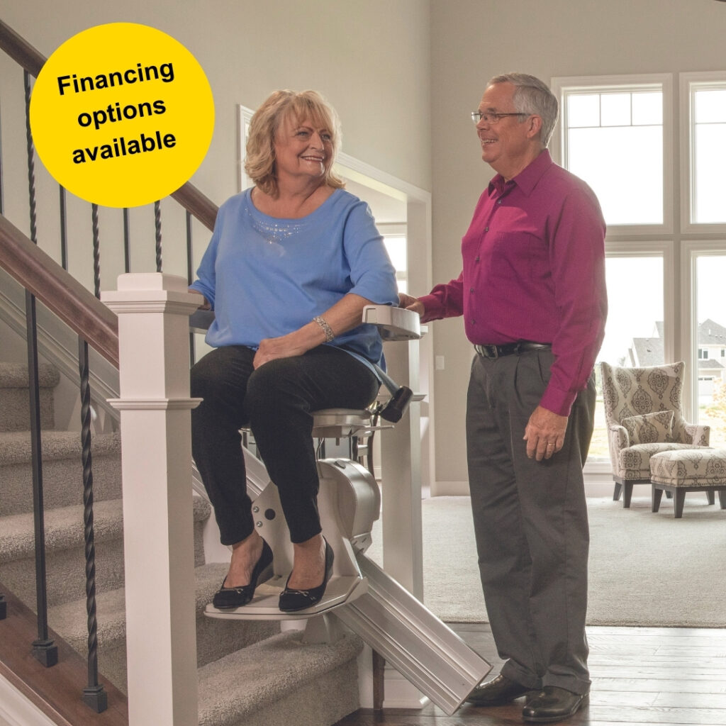 Senior couple pictured with woman seated on stairlift chair and man standing beside her at the bottom of the staircase. A round yellow sticker is in the top left corner and reads financing options available. 