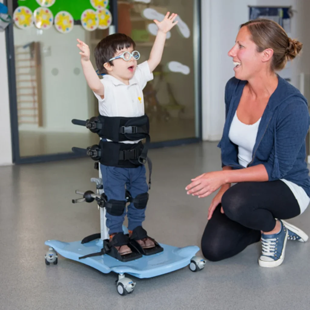 Young boy with glasses arms stretched upwards using the R82 Meerkat Stander. A woman is kneeling to his right. 