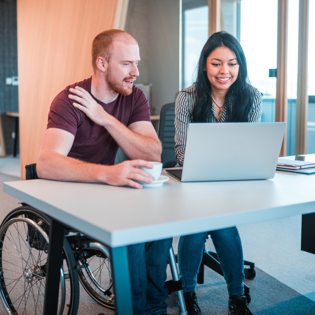 Male wheelchair user seated with a young woman at a desk looking at an open laptop.