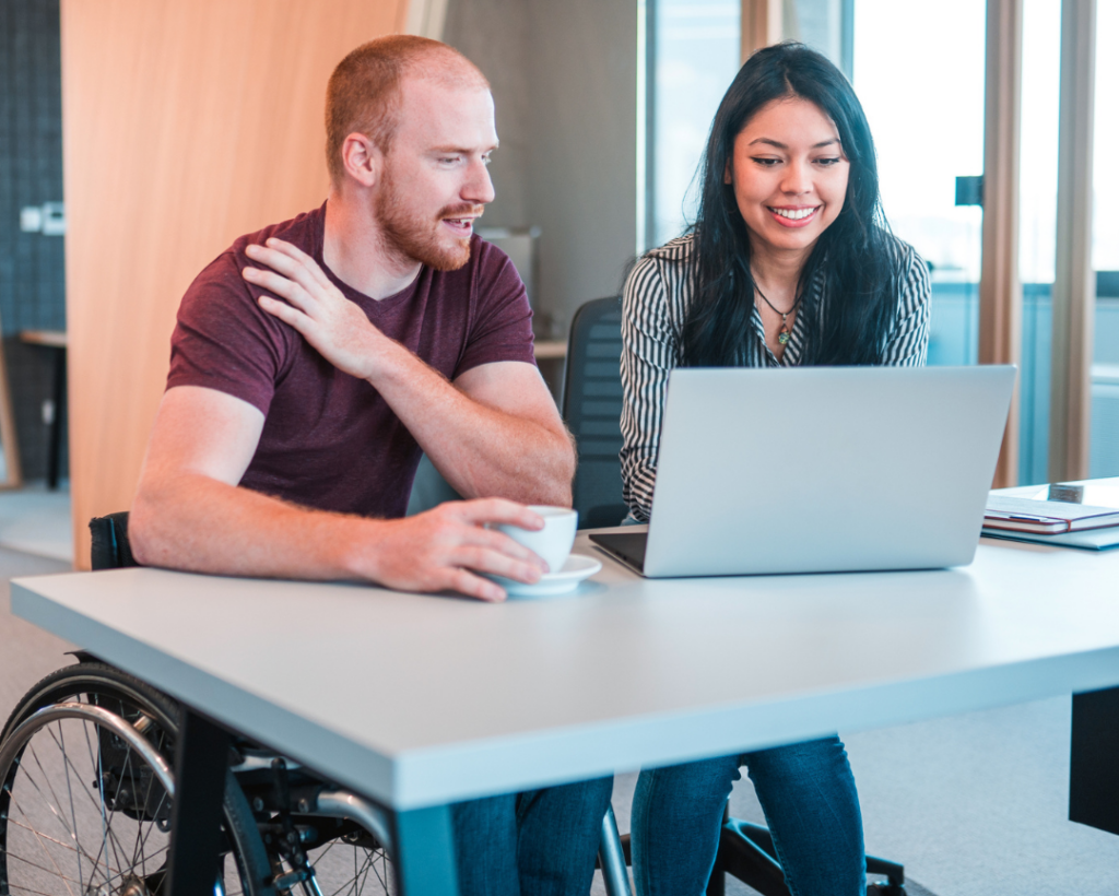 Male wheelchair user seated with a young woman at a desk looking at an open laptop.